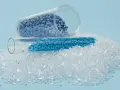 Granulat plastiku w probówkach i rozsypany na jasnoniebieskim tle.