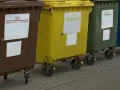 Rząd kolorowych pojemników na śmieci: brązowy (bio), żółty (metale i tworzywa sztuczne), zielony (szkło), niebieski (papier i tektura).