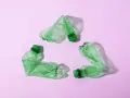 Trzy zielone, zgniecione butelki tworzą symbol recyklingu na różowym tle.