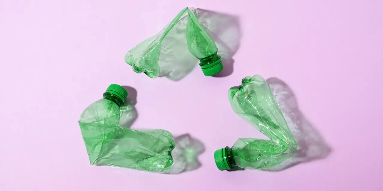Trzy zielone, zgniecione butelki tworzą symbol recyklingu na różowym tle.