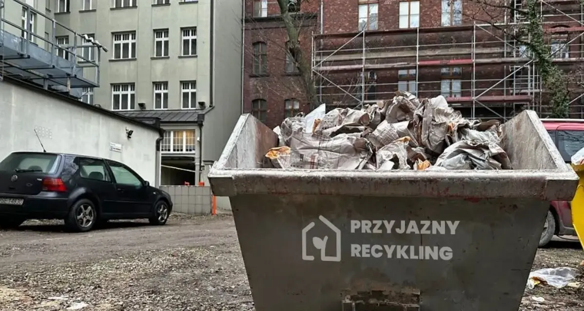Kontener na śmieci z napisem "Przyjazny Recykling" wypełniony papierem. W tle budynki i czarny samochód. Data: 17 09 04.