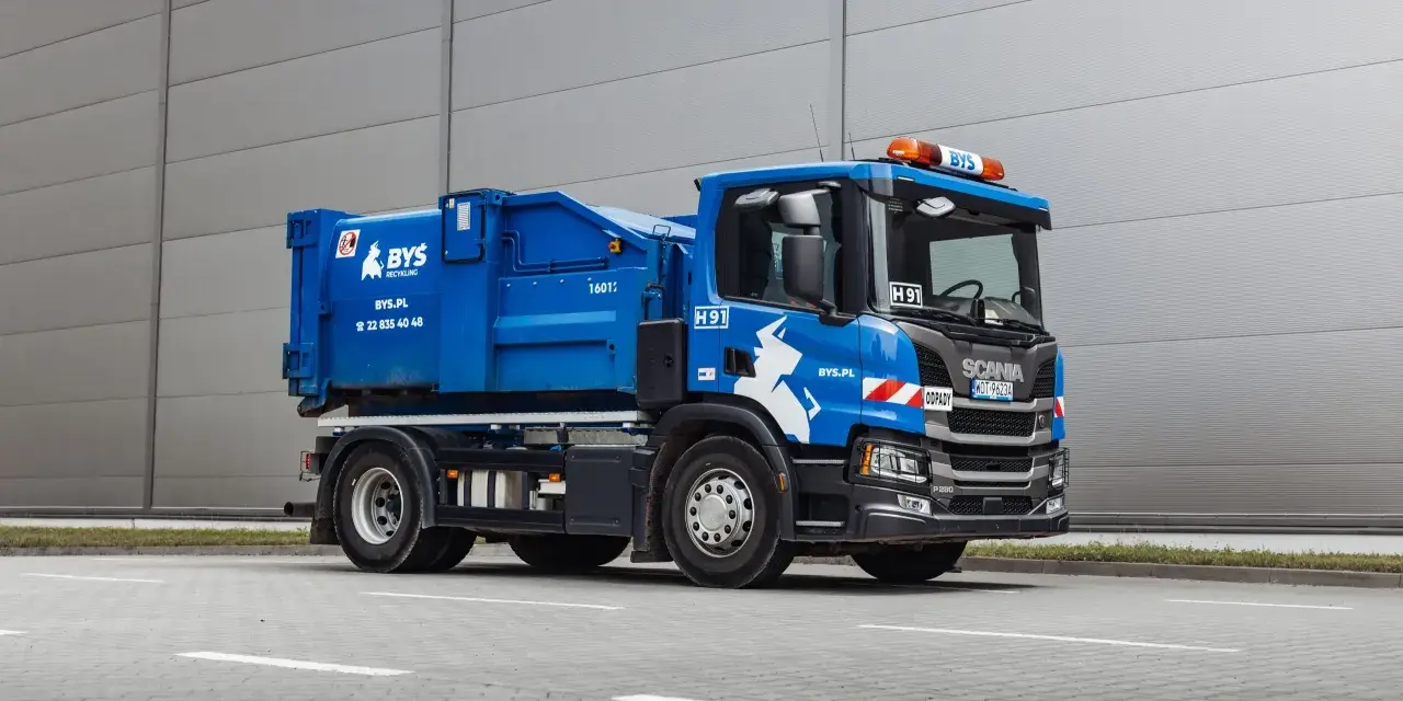 Niebieska śmieciarka Scania z napisem "ODPADY" i logo BYS. Pojazd gotowy do odbioru gruzu, z kodem odpadu na tablicy.