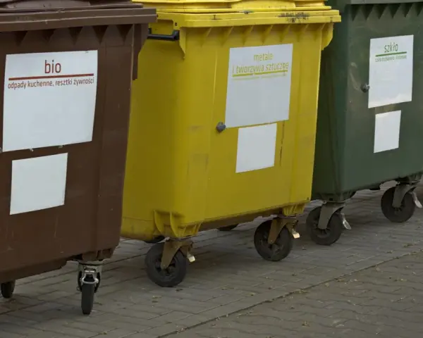 Rząd kolorowych pojemników na śmieci: brązowy (bio), żółty (metale i tworzywa sztuczne), zielony (szkło), niebieski (papier i tektura).