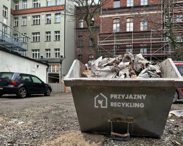 Kontener na śmieci z napisem "Przyjazny Recykling" wypełniony papierem. W tle budynki i czarny samochód. Data: 17 09 04.