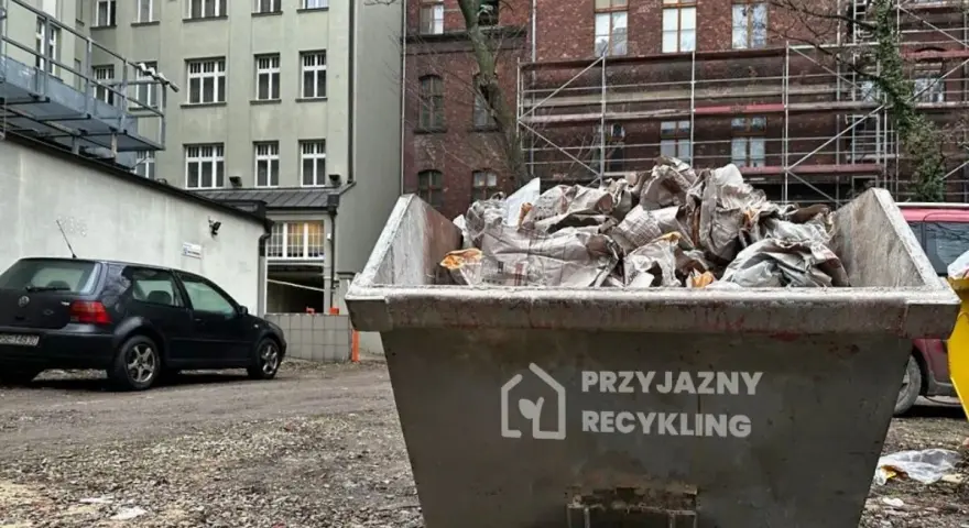 Kontener na śmieci z napisem "Przyjazny Recykling" wypełniony papierem. W tle budynki i czarny samochód. Data: 17 09 04.