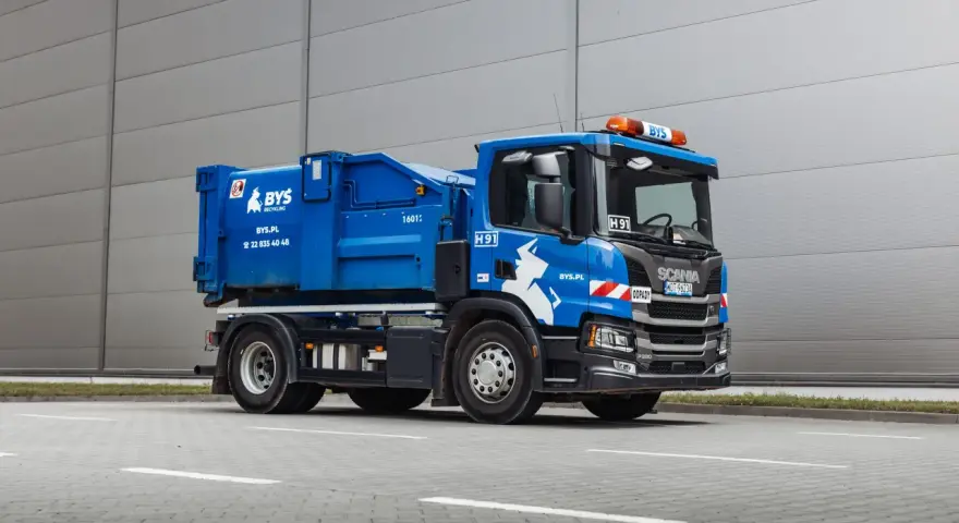 Niebieska śmieciarka Scania z napisem "ODPADY" i logo BYS. Pojazd gotowy do odbioru gruzu, z kodem odpadu na tablicy.