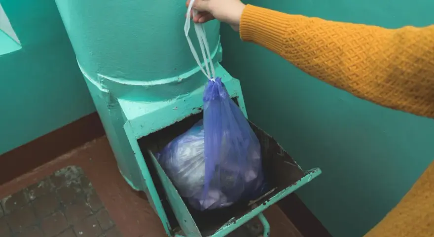 Ręka w żółtym swetrze wrzuca worek ze śmieciami do zsypu w bloku.